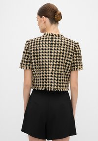 Svart och tan rutigt kort jacka med korta ärmar, fransade kanter och en texturerad tyg. Tillsammans med svarta shorts.