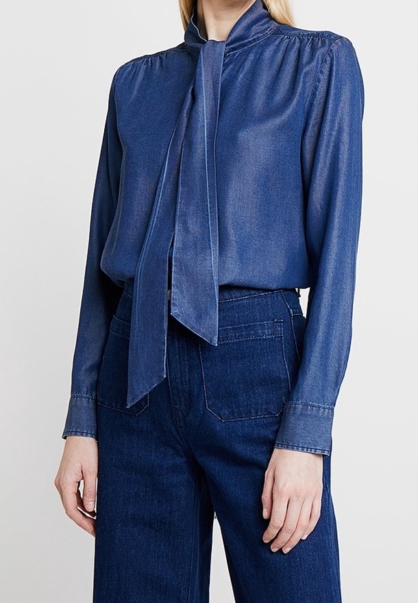 Eine Frau trägt eine langärmelige blaue Denim-Bluse mit einem vorn gebundenen Kragen, kombiniert mit dunkelblauen hochgeschnittenen Denim-Jeans.