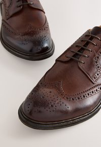 Brune læderbrogues med spids tå, perforerede detaljer og snøredesign med kontrasterende mørke accenter på sålen.