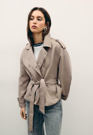 Mlada ženska, oblečena v bež trenč plašč z pasom, velike srebrne uhane, sive kavbojke in belo majico, stoji pred enobarvnim svetlim ozadjem.