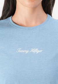 Šviesiai mėlynas medvilninis marškinėliai su apvaliu kaklu ir trumpomis rankovėmis, ant priekio puikuojasi baltas kursyvu rašytas "Tommy Hilfiger" logotipas.