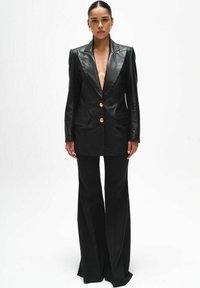 Kobieta stojąca prosto, ubrana w czarną skórzaną marynarkę z złotymi guzikami oraz szerokie czarne spodnie na tle jednolitej białej ściany.