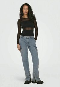Top preto em malha de mangas longas com seções transparentes, combinado com calças de carga azul claro com bolsos laterais e fechos, usado com calçado preto.