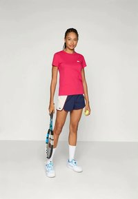 Ružový tričko s okrúhlym výstrihom skombinované s námorníckymi a krémovými športovými šortkami. Model drží tenisovú raketu a loptičku, má na sebe biele tenisky s modrými akcentmi.