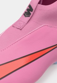 Ružičaste nogometne kopačke s glatkom, teksturiranom površinom. Imaju hrabar crveni swoosh dizajn i crni logo blizu uzice.