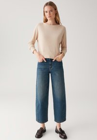 Maglione beige, strutturato, con maniche lunghe e taglio cropped, abbinato a jeans in denim blu a vita alta e gamba larga, con mocassini scuri.