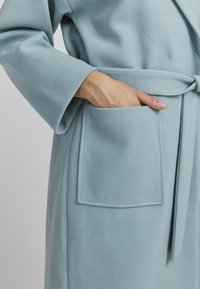 Robe bleu clair avec une finition texturée, dotée d'une taille ceinturée et d'une poche à rabat sur le côté gauche. Manches longues avec une coupe décontractée.