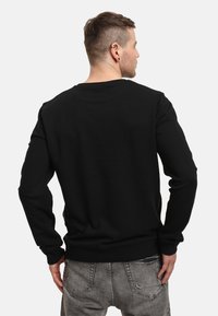 Uomo visto di spalle che indossa una camicia nera a maniche lunghe e jeans grigi, in piedi con le mani vicino alle tasche su uno sfondo neutro.