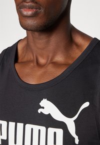Svart tanktopp med rund hals, med en fremtredende hvit Puma-logo. Stoffet ser glatt og figurnært ut, og viser detaljer på skuldrene og nakken.