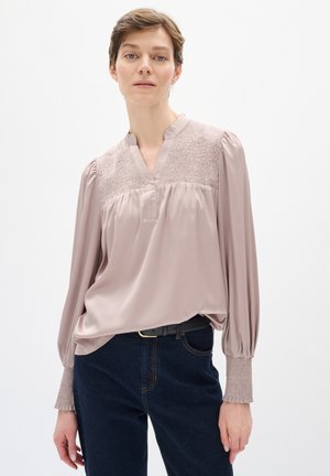 Blushfarbene Seidenbluse mit tiefem V-Ausschnitt, gerafften Details an den Schultern, langen Ärmeln mit gerafften Manschetten, kombiniert mit dunkelblauen Jeans.