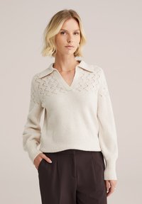 Pleteni pulover v kremni barvi z V-izrezom in srajčnim dizajnom. Ima vzorec z odprtimi deli na ramenih in rebraste manšete, kombiniran s temnimi hlačami.