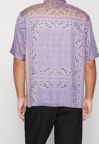 Krátký rukáv košile ze světle fialové látky, zdobená krajkovým vzorem a květinovými akcenty, s uvolněným střihem a jemným leskem.