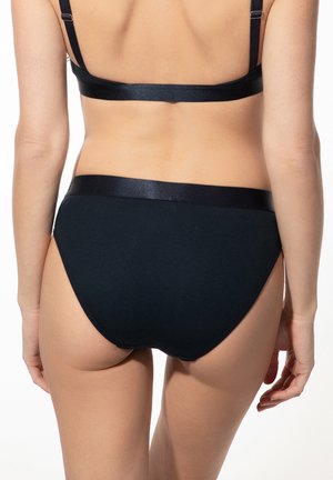 Woman wearing black satin-trimmed underwear and matching bra, shown from the back against a plain light background.