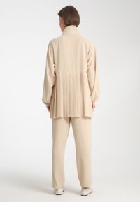 Giacca beige a pieghe con colletto alto, maniche lunghe e un taglio morbido, abbinata a pantaloni beige coordinati e scarpe di colore chiaro.