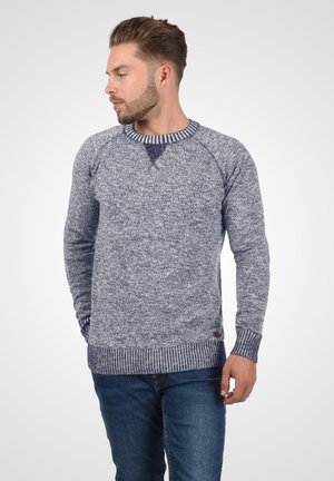 Mężczyzna z krótkimi włosami i brodą, ubrany w niebieski melanżowy sweter i niebieskie dżinsy, patrzy w lewo, trzymając ręce blisko kieszeni.