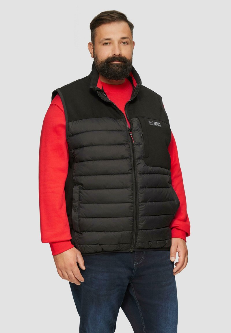 Mann mit Bart, der eine schwarze gepolsterte Weste über einem roten Langarmshirt und blauen Jeans trägt, stehend vor einem schlichten grauen Hintergrund.