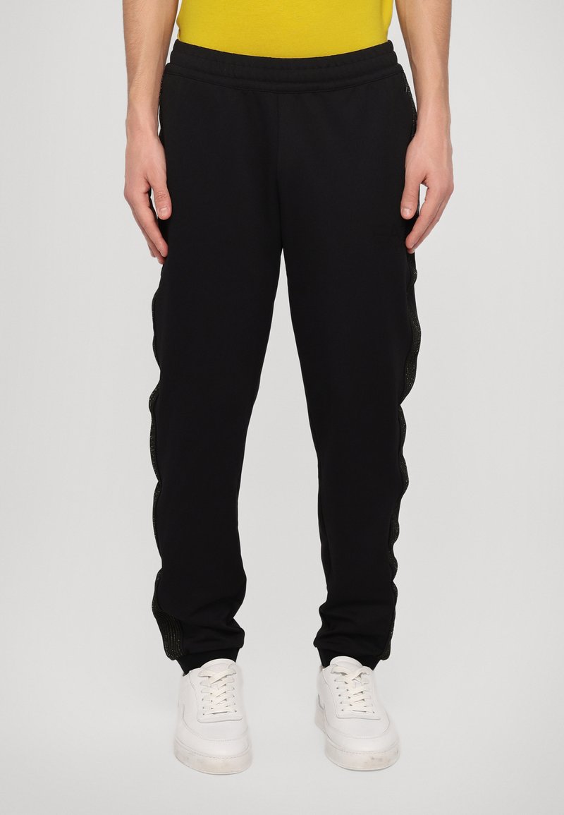 Zwarte trainingsbroek met een gestructureerd, golvend zijpaneel en elastische tailleband, gecombineerd met witte sneakers en een gele shirt.
