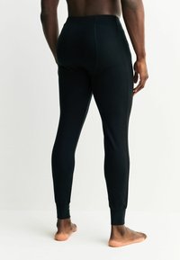 Čierne legíny z elastickej tkaniny, so priliehavým strihom a rebrovanými manžetami pri členkoch, na neobutých nohách z pohľadu zozadu.