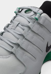 Κοντινό πλάνο γκρι αθλητικού παπουτσιού Nike με επάνω μέρος από δίχτυ, μοναδικούς βρόχους κορδονιών, μαύρο λογότυπο swoosh και πράσινες λεπτομέρειες στη γλώσσα και τη σόλα.