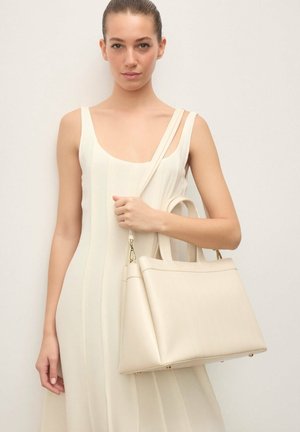 Joven mujer con vestido blanco sin mangas sosteniendo un gran bolso de cuero crema con correa para el hombro sobre un fondo liso.