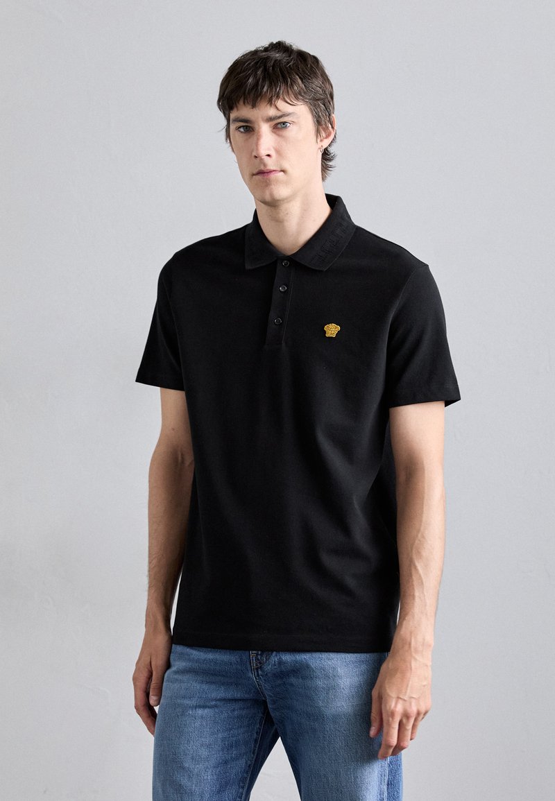 Polo noir à manches courtes avec un col structuré, patte de boutonnage à trois boutons et petit logo doré sur la poitrine. Associé à un jean bleu.
