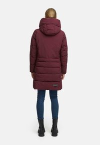 Bordeauxkleurige pufferjas met een hoge kraag en capuchon, voorzien van een gestikt ontwerp. Draagt bovenop een blauwe spijkerbroek en zwarte laarzen. Achteraanzicht weergegeven.