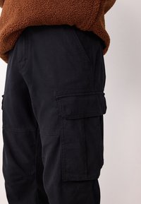 Pantaloni cargo in cotone blu scuro con tasche laterali, cuciture visibili e vestibilità comoda, abbinati a un maglione marrone in pile con trama.