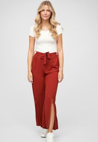Pantaloni larghi rossi in tessuto texture con cintura annodata e aperture frontali, abbinati a una maglietta bianca a maniche corte e sneaker bianche.