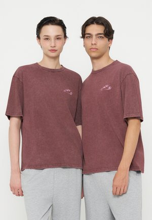 Deux jeunes adultes portant des T-shirts bordeaux délavés assortis avec de petits logos et des pantalons gris clair, debout côte à côte.