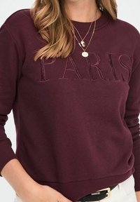 Felpa bordeaux con "PARIGI" ricamato in una tonalità abbinata, dotata di scollatura a girocollo e polsini e orlo a costine. Texture liscia.