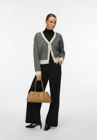 Svart och vit mönstrad cardigan med en beige kant, bärs över en svart polotröja. Matchad med svarta vida byxor och en brun handväska.