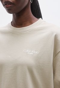 Tricou bej din bumbac cu decolteu rotund și mâneci scurte, având logo-ul brodat alb „Calvin Klein JEANS” pe piept.