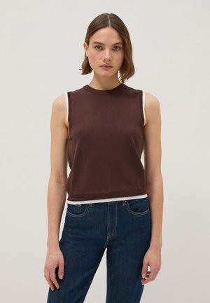 Jonge vrouw met kort bruin haar die een mouwloze donkerbruine top met witte bies en donkerblauwe spijkerbroek draagt, staand tegen een effen achtergrond.