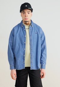 Blauwe denim shirt met een knoopsluiting en puntige kraag, gedragen over een lichtgroen t-shirt. Zwarte shorts en een zwarte pet maken de outfit compleet.