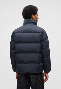 Casaco acolchoado azul marinho com gola alta e design acolchoado, apresentando secções horizontais grossas e acabamento mate.