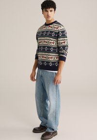 Suéter de punto en color crema con acentos en azul marino, que presenta un patrón geométrico en rojo, verde y azul. Combinado con jeans de color azul claro y zapatos marrones.