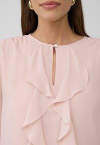 Sieviete valkā gaiši rozā blūzi ar vienu augšējo pogu un plūstošu volānu dekoru pa priekšu, kombinējot to ar zelta riņķa auskariem.