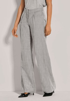 Hellegraue weitgeschnittene Hose aus strukturiertem Stoff, mit vorderen Falten und einem glatten Bund. Kombiniert mit schwarzen spitzen High-Heels.