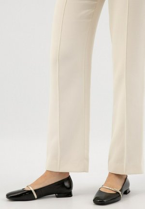Black patent leather flats with a square toe and small heel, featuring a thin, metallic strap across the instep. Worn with cream-colored pants.