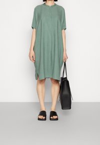 Volné zelené šaty s krátkými rukávy, kulatým límcem a bočními rozparky. Spárováno s černými pantofli a černou taškou. Hladká textura.