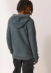 Hoodie tricoté vert foncé avec des détails en côtes, en tissu texturé et une coupe décontractée. Doté d'une capuche et de larges poignets, porté avec un pantalon cargo noir.