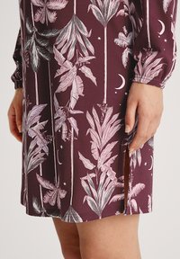 Levendige bordeaux jurk met tropische bladmotieven in roze en groen, lange mouwen met geplooide manchetten en een zijsplits.