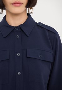 Navy blue shirt featuring a collar, button-up front, two chest pockets, and shoulder epaulettes made of smooth fabric. Large silver earrings visible.
