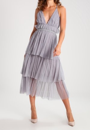 Donna che indossa un abito midi grigio senza maniche, a balze e plissettato con dettagli argentati e tacchi alti color nude, in piedi davanti a uno sfondo uniforme.