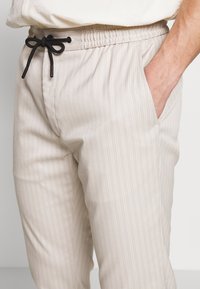 Beige randiga byxor med elastisk midja och svart dragsko, med sidofickor och en mjuk tygtextur.