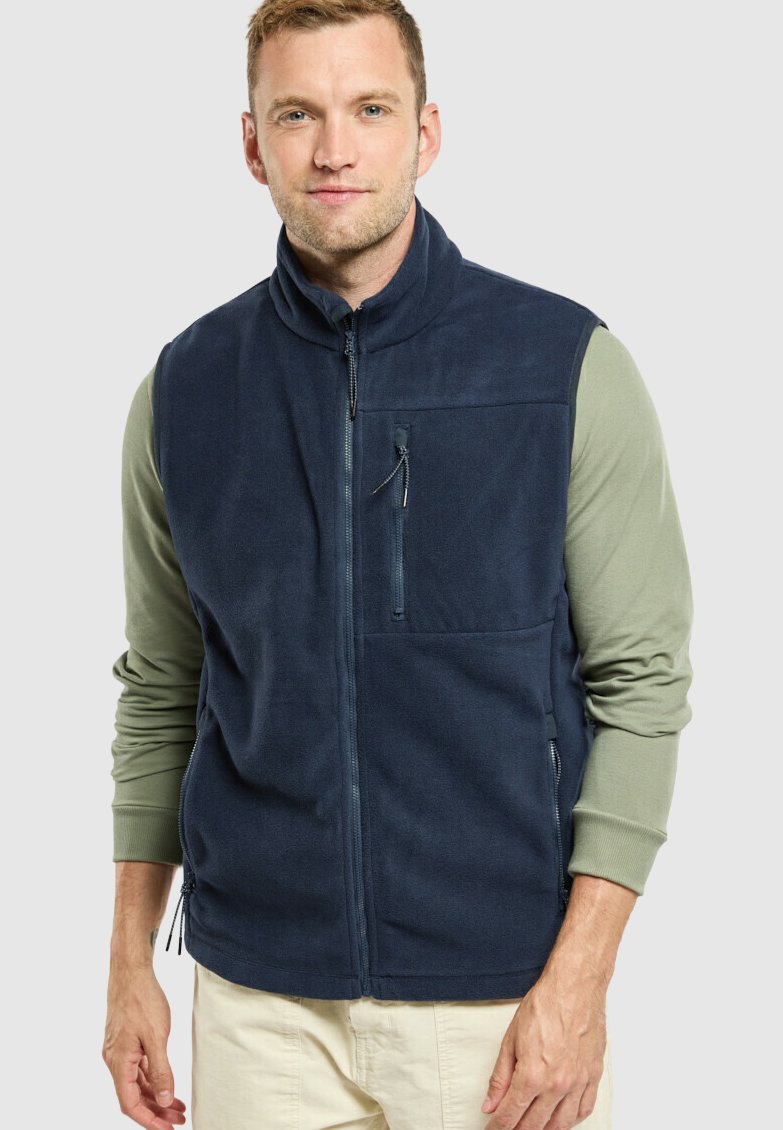 Gilet en polaire marine avec col haut, poches zippées et texture lisse. Porté par-dessus une chemise à manches longues vert clair. Fermeture éclair à l'avant.