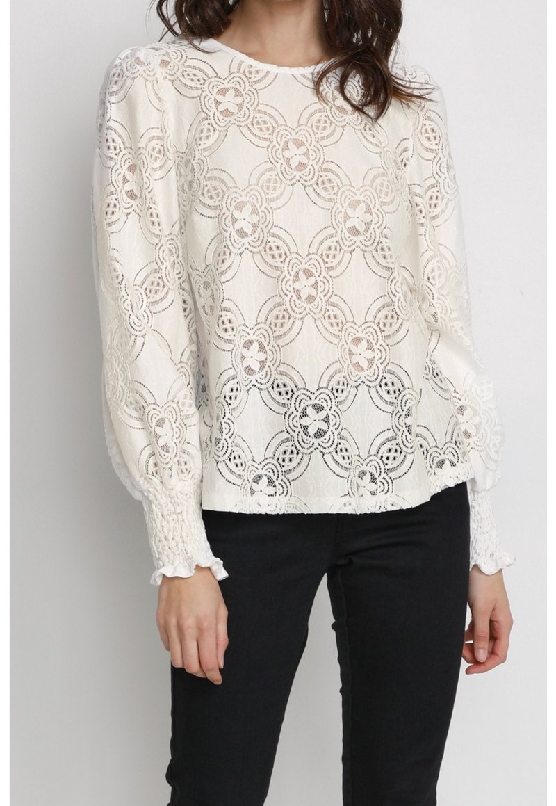 Blouse blanche en dentelle à manches longues avec motifs floraux circulaires et poignets froncés, portée avec un pantalon noir sur une personne debout.