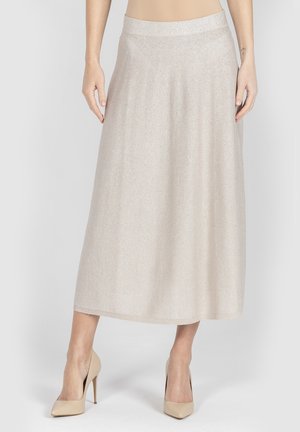 Kobieta w beżowej, błyszczącej spódnicy midi, w połączeniu z pasującymi beżowymi szpilkami, stoi na jednolitym tle.