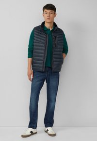 Marinefarbener Steppweste mit horizontalem Quiltmuster, Frontreißverschluss und Seitentaschen, über einem grünen Poloshirt, kombiniert mit blauen Jeans und weißen Turnschuhen.