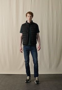 Sort polstret vest over en mørk t-shirt, blå jeans og sorte sneakers. Vestens lukning er med lynlås, og den har en kviltet tekstur.